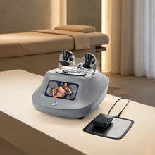 A tecar therapy beauty device with dual black applicator heads sits on a beige table in a spa-like room, with a black charging pad and cable beside it and a massage table in the background.